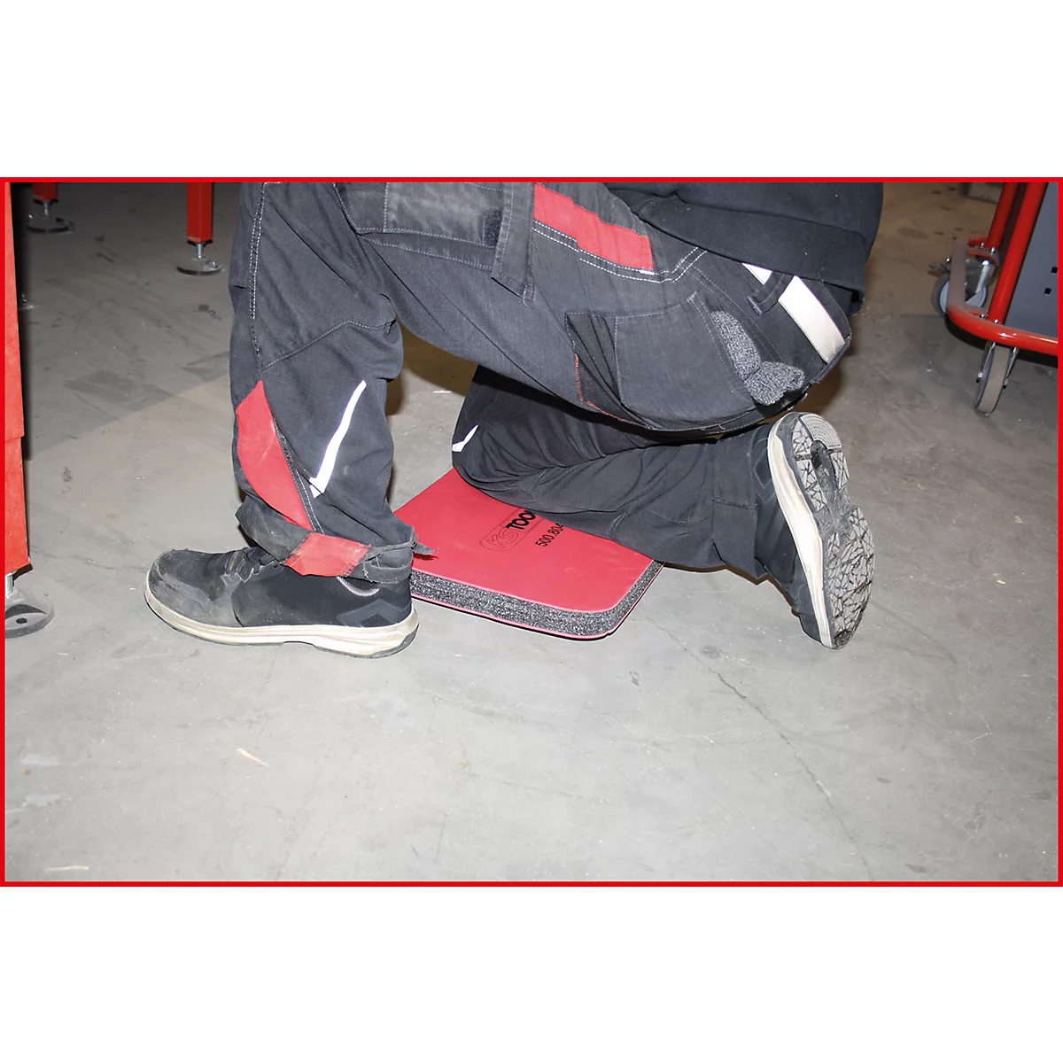 Arbeiter kniet auf einer roten Knieschutzmatte, schwarze Hose mit roten Akzenten, schwarze Schuhe.